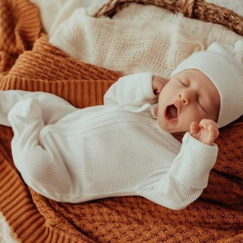 Yawning baby in a white my first outfit with matching beanie