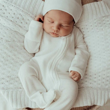 Baby lying on a white blanket in a my first outfit and matching beanie also in white