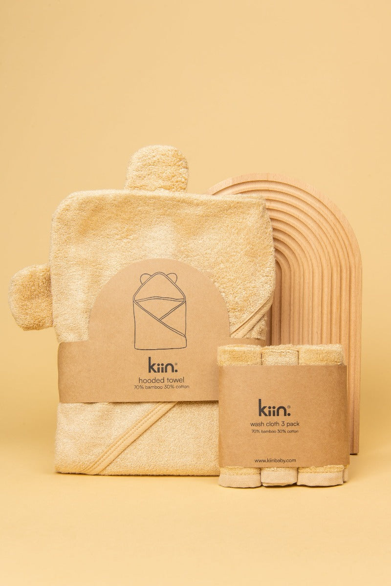 Oat Hooded Towel and matching washcloths in packaging standing up against a wooden rainbow