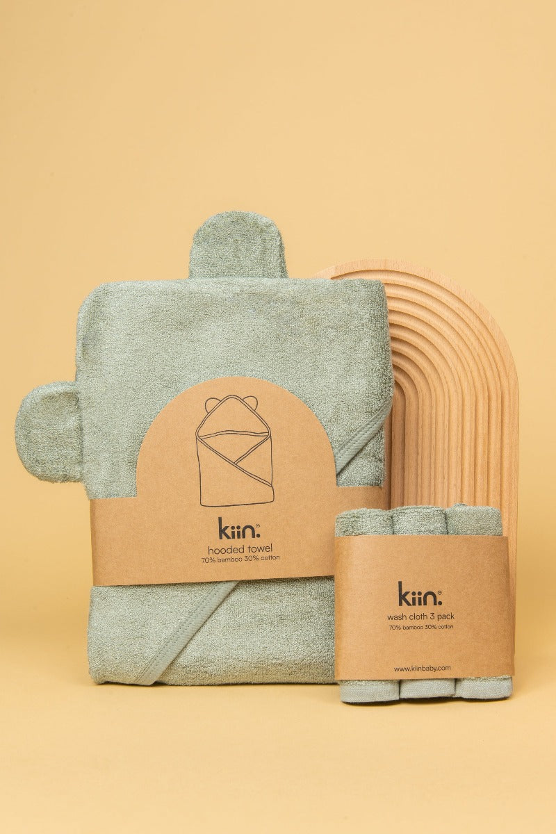 Sage Hooded Towel and matching washcloth in packaging standing up against a wooden rainbow
